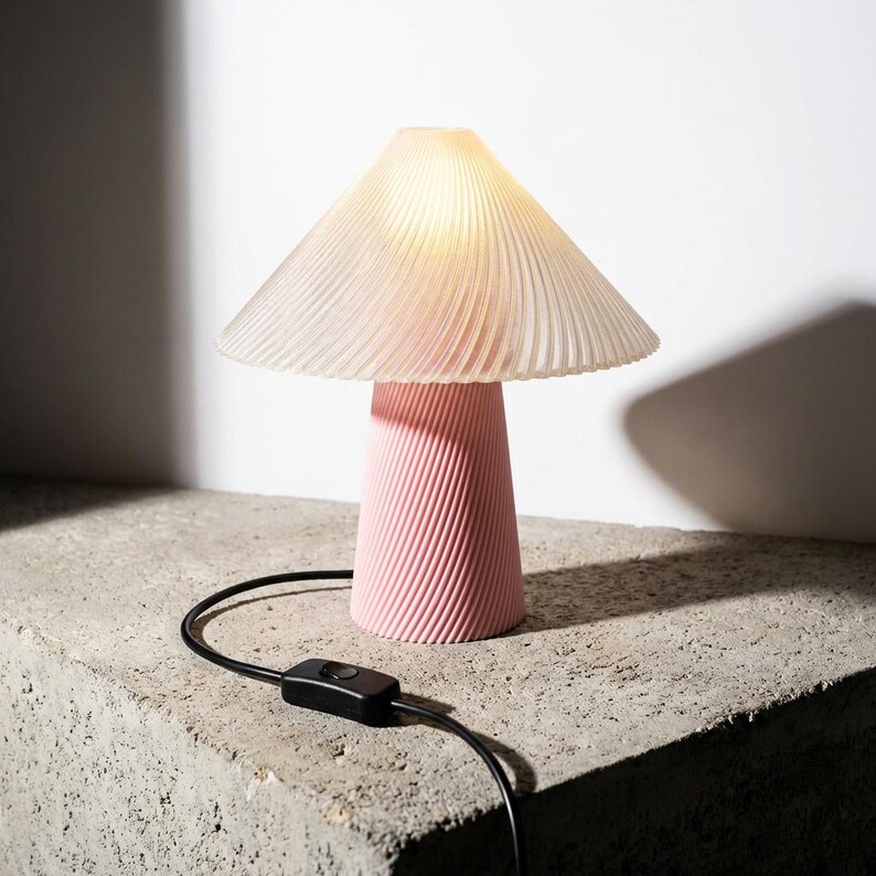 May include: A pink mushroom-shaped table lamp with a ribbed base and a pleated, translucent lampshade. The lamp is turned on, emitting a soft glow. A black power cord with an on/off switch extends from the base. The lamp sits on a concrete surface.