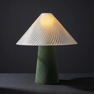 May include: A modern table lamp with a white, pleated lampshade and a green, textured base. The lamp's conical base has a spiral design. The lampshade is lit, casting a warm glow. The lamp is on a dark surface.