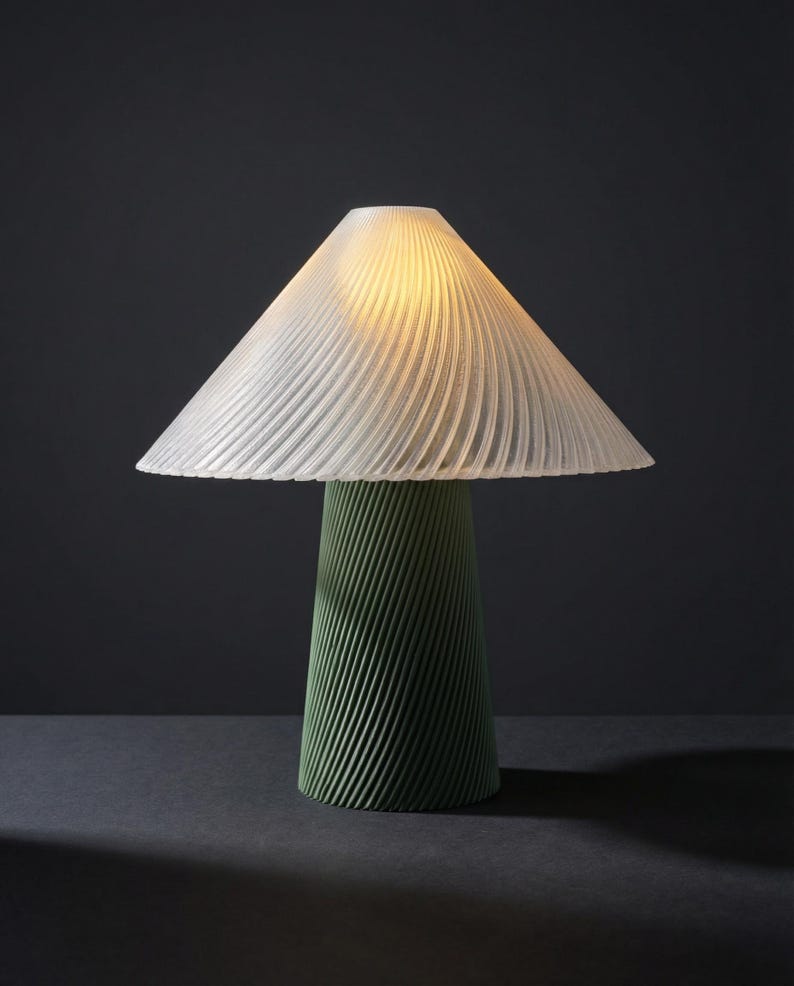May include: A modern table lamp with a white, pleated lampshade and a green, textured base. The lamp's conical base has a spiral design. The lampshade is lit, casting a warm glow. The lamp is on a dark surface.