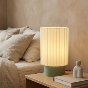 May include: A modern table lamp with a pleated white shade and a light green base. The lamp sits on a wooden nightstand next to a small white vase and a book. The lamp is turned on, illuminating the room.
