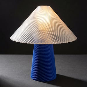 May include: A modern table lamp with a blue base and a clear, ribbed lampshade. The base has a conical shape with diagonal grooves. The lampshade is a translucent, inverted cone shape, and the lamp is lit from within, casting a warm glow.