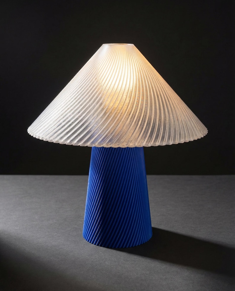 May include: A modern table lamp with a blue base and a clear, ribbed lampshade. The base has a conical shape with diagonal grooves. The lampshade is a translucent, inverted cone shape, and the lamp is lit from within, casting a warm glow.