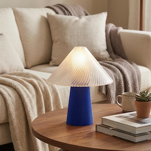 May include: A modern table lamp with a blue base and a white, pleated lampshade. The lamp is on a round, wooden table, with a neutral-toned sofa and throw pillows in the background. A small potted plant and books are also on the table.