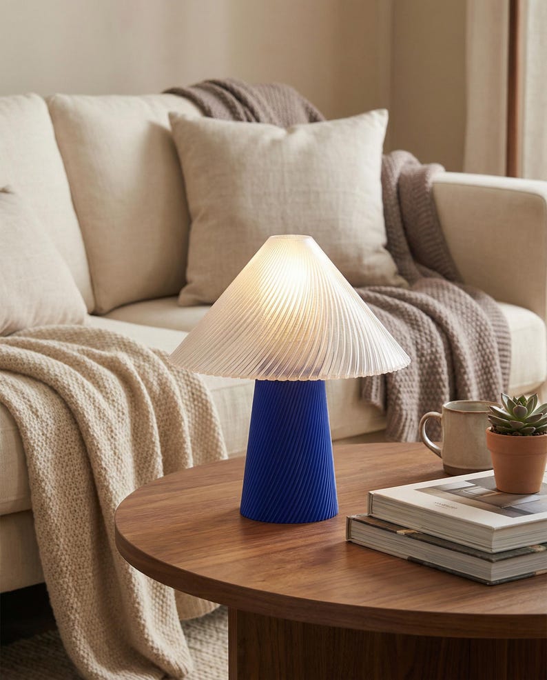 May include: A modern table lamp with a blue base and a white, pleated lampshade. The lamp is on a round, wooden table, with a neutral-toned sofa and throw pillows in the background. A small potted plant and books are also on the table.