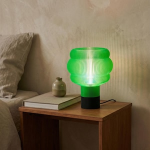 May include: A table lamp featuring a vibrant green, ribbed, translucent shade with a rounded, bulbous silhouette. The lamp has a solid black cylindrical base and is illuminated from within, casting a soft glow through the layered structure.