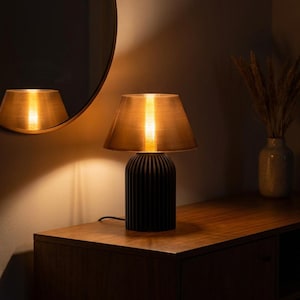 May include: A modern table lamp with a black, ribbed base and a gold-toned lampshade. The lamp is on a wooden surface, with a mirror reflecting the light. The warm light creates a cozy atmosphere. Home decor.