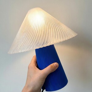 May include: A modern table lamp with a pleated white lampshade and a vibrant blue base. The lamp is turned on, illuminating the room with a soft glow. The design features a unique, textured surface.