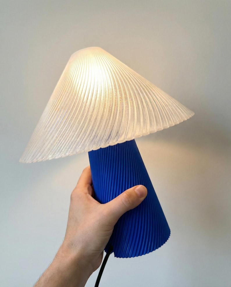May include: A modern table lamp with a pleated white lampshade and a vibrant blue base. The lamp is turned on, illuminating the room with a soft glow. The design features a unique, textured surface.