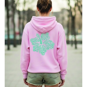 以下が含まれることがあります： 背中に大きく詳細な、明るい緑色のハイビスカスの花の模様が施された、明るいピンク色のパーカー。 パーカーにはフードと長袖が付いています。 モデルはデニムショーツを着用しています。