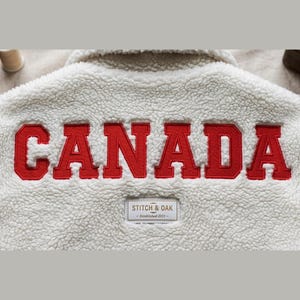 May include: A white sherpa fleece jacket featuring the word CANADA in large red block letters across the back. Below the text is a rectangular white patch with a brown border that reads STITCH & OAK and Established 2023 in brown text.