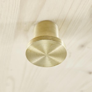 May include: A close-up shot of a brass cabinet knob. The knob has a cylindrical body and a circular, ridged base. It is set against a light-coloured wooden surface, highlighting its metallic sheen and detailed design.