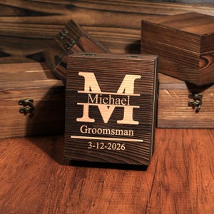 May include: A dark brown wooden box with the letter "M" and the name "Michael" engraved on the front. Below the name are the Roman numerals "IVI", the word "Groomsman", and the date "3-12-2026". Other wooden boxes are in the background.