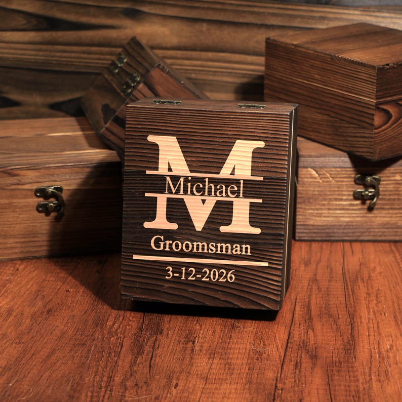 May include: A dark brown wooden box with the letter "M" and the name "Michael" engraved on the front. Below the name are the Roman numerals "IVI", the word "Groomsman", and the date "3-12-2026". Other wooden boxes are in the background.