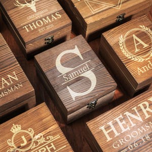 May include: A collection of personalized wooden boxes with various engraved designs. The boxes are brown with metal clasps. Some boxes feature names like "Thomas" and "Samuel", initials, and the text "Groomsman".