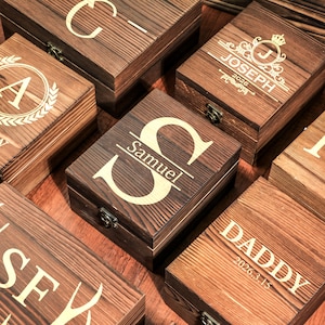 May include: A collection of personalized wooden boxes with hinged lids. Each box is stained a dark brown and features a unique engraved design, including initials, names, and dates. The boxes are closed with metal clasps.