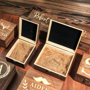 May include: Several open wooden boxes with personalized engravings. The boxes are filled with straw and have black velvet lining. One box lid reads "Robert" and "SMAN 15". Other boxes have decorative elements and names.