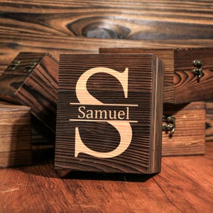 May include: A wooden box with a dark brown finish, featuring a large, light-colored letter "S" and the name "Samuel" engraved below. Other wooden boxes are visible in the background, all with a similar dark brown finish.