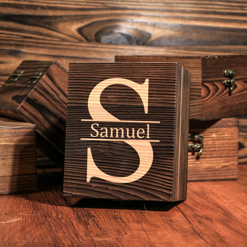 May include: A wooden box with a dark brown finish, featuring a large, light-colored letter "S" and the name "Samuel" engraved below. Other wooden boxes are visible in the background, all with a similar dark brown finish.