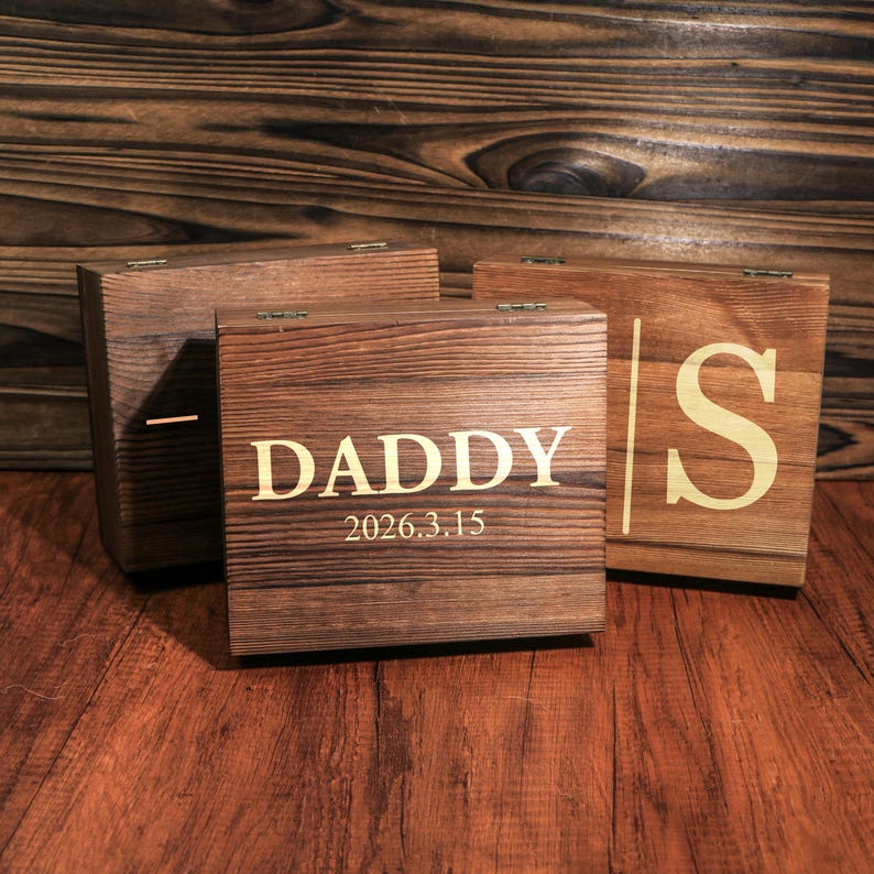 May include: Four wooden boxes with hinged lids. The front box is engraved with the word "DADDY" and the date "2026.3.15" in a gold color. The other boxes have a similar wood grain and gold-colored engravings.