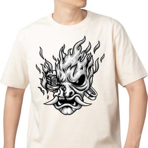 May include: A cream-colored short-sleeve t-shirt featuring a black and white graphic print of a stylized oni mask combined with mechanical elements and flames. The design includes a robotic eye, sharp fangs, and intricate line work details.