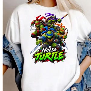 May include: A white short-sleeved t-shirt featuring a graphic print of four green ninja turtles with colored masks and their respective weapons. Below the characters, the text NINJA TURTLE is printed in a bold, stylized green and black font.