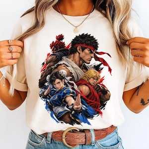 Puede incluir: Una camiseta de color crema con un estampado gráfico de cuatro personajes de la serie de videojuegos Street Fighter, incluidos Ryu, Ken, Chun-Li y Akuma, representados en una composición dinámica y superpuesta con llamativos acentos de tinta roja y azul.