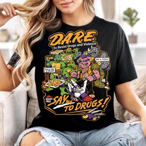Op de afbeelding: Een zwart t-shirt met een grafische print met de tekst D.A.R.E. To Resist Drugs and Violence. Het ontwerp bevat stripfiguren die pizza eten, tekstballonnen met de tekst Which one is drugs? en Say a pizza!, en de tekst Say no to drugs!
