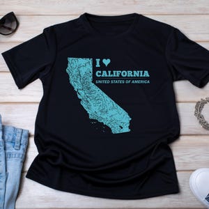 Può includere: Una t-shirt nera a maniche corte con una grafica azzurra dello stato della California con una texture cartografica dettagliata. Il design include il testo I heart CALIFORNIA UNITED STATES OF AMERICA stampato in un carattere sans-serif azzurro.