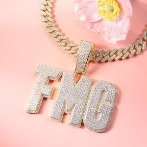 May include: A gold-toned, iced-out necklace with a large pendant spelling out "FMG" in sparkling, clear stones. The chain is a thick, linked design. The background is a soft pink with a flower.