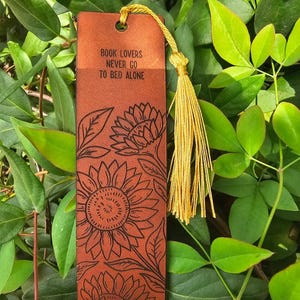 May include: A brown leather bookmark with the text "BOOK LOVERS NEVER GO TO BED ALONE" and a sunflower design. The bookmark has a gold tassel attached to the top. The background is green foliage.
