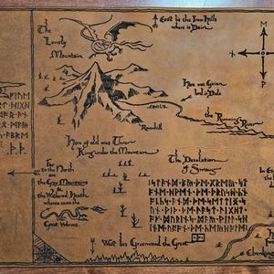 May include: A detailed map with hand-drawn illustrations and text, featuring mountains, a dragon, and a compass. The map is in shades of brown and black, with text in a stylized font. The map includes the text "The Lonely Mountain" and "The Desolation of Smaug".