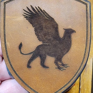 May include: A shield-shaped leather patch with a detailed griffin design. The griffin, a mythical creature with an eagle's head and wings and a lion's body, is etched in dark brown against a lighter brown background. The patch has a dark brown border.
