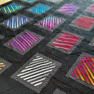 Op de afbeelding: Close-up van een handgemaakte quilt met een zwarte achtergrond en ruitvormige patronen. Elke ruit heeft kleurrijke, diagonale strepen in tinten blauw, paars, rood en geel. De quilt heeft een spiraalvormig quiltontwerp.