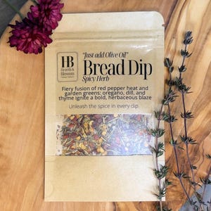 May include: A package of "Bread Dip Spicy Herb" with a clear window showing the spice blend. The package is tan with the text "Just add Olive Oil" and "Hearth & Blossom". The spice blend includes red pepper flakes and green herbs. Purple flowers and a sprig of thyme are in the background.