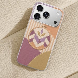 Op de afbeelding: Een doorzichtig smartphonehoesje met een print van een ronde borduurring met een geometrisch quiltpatroon in tinten paars, beige en crème, geaccentueerd door een klein bladmotief, tegen een achtergrond van gelaagde stoftexturen.