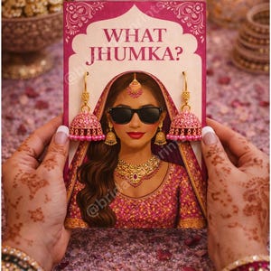 May include: A pink and gold illustration of a woman wearing sunglasses and traditional Indian jewelry, holding a sign that reads "WHAT JHUMKA?". The woman is wearing a pink and gold outfit. The image is held by hands with henna tattoos.