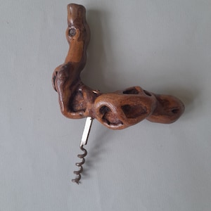 May include: A handcrafted wooden corkscrew with a unique, organic shape. The corkscrew features a natural wood grain and a metal worm. The handle has a distinctive, textured design with several holes.