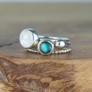 May include: A set of three silver rings with different stones. The top ring features a large, round, white moonstone. The middle ring has a smaller, round, green labradorite stone. The bottom ring has a silver bead.