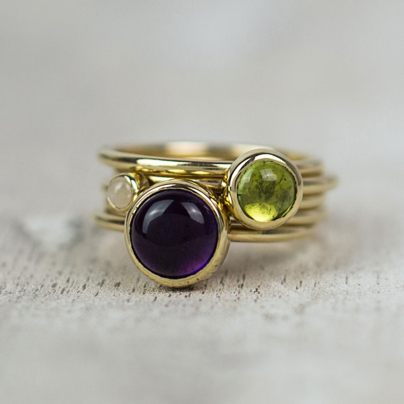May include: A gold ring with three stacked bands and three gemstones. The gemstones are a purple amethyst, a green peridot, and a white diamond.
