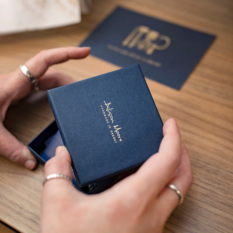 May include: A navy blue jewelry box with gold lettering that reads "Alison Moore Handmade in Orkney".