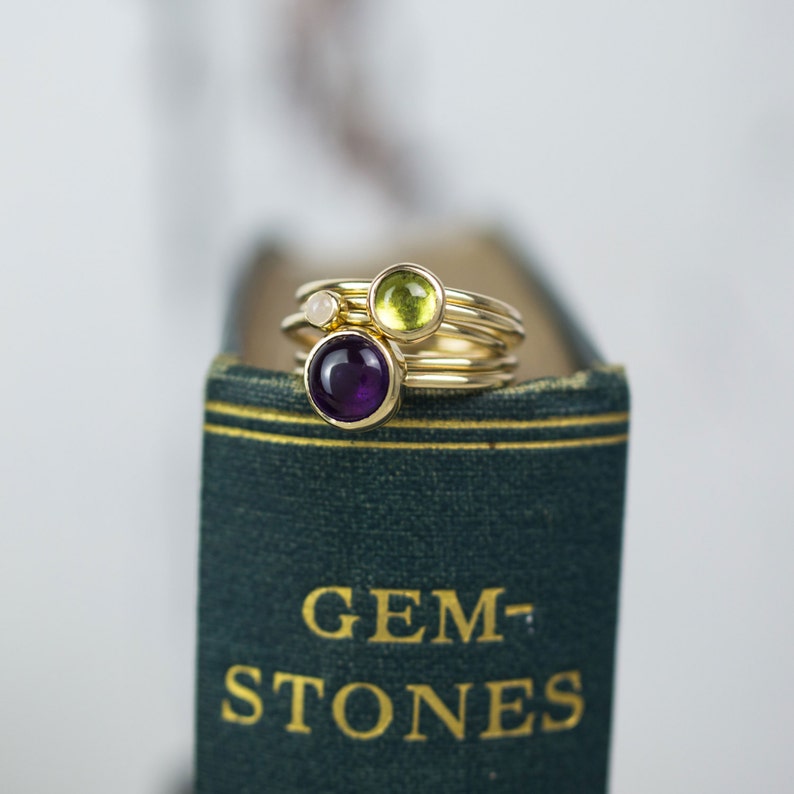 May include: A set of three gold rings with different colored gemstones. The rings are stacked on top of each other. The top ring has a green gemstone, the middle ring has a white gemstone, and the bottom ring has a purple gemstone. The rings are sitting on a green book with the words "GEM-STONES" printed on the cover.
