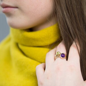 May include: A close-up of a hand wearing a stack of gold rings with purple and green gemstones.