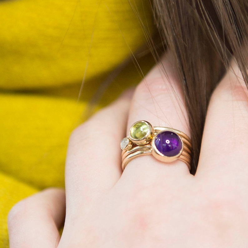 May include: A set of three gold rings with gemstones. The rings feature a purple amethyst, a green peridot, and a clear diamond. The rings are stacked on a finger.