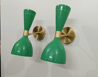May include: Two green wall sconces with a brass finish. The sconces have a conical shade with small holes near the top and a cylindrical neck. The wall mounts are circular and made of brass. The sconces are mounted on a white wall.