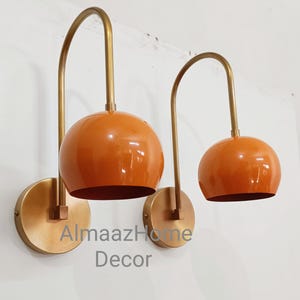 Op de afbeelding: Twee oranje globevormige wandlampen met messingkleurige gebogen armen en ronde achterplaten. De lampen zijn op een witte muur gemonteerd. De tekst "AlmaazHome Decor" staat op de muur.