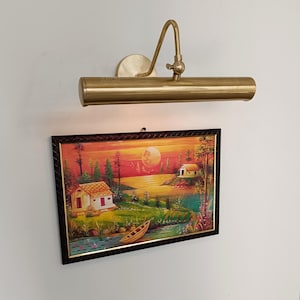 May include: A brass picture light above a framed landscape painting. The painting shows a sunset scene with a river, houses, and trees. The frame is dark brown with a gold inner edge.