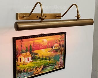 May include: A brass picture light fixture mounted above a framed landscape painting. The painting depicts a sunset scene with a house, trees, and a boat. The light fixture has a rectangular base and a curved arm.