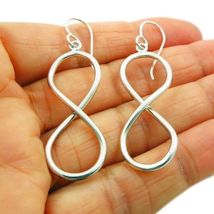 May include: A pair of silver-colored infinity symbol earrings. The earrings are made of a smooth, shiny metal and feature a simple, elegant design. The infinity symbol is a continuous loop, creating a modern and minimalist look. The earrings are shown against a skin-toned background.