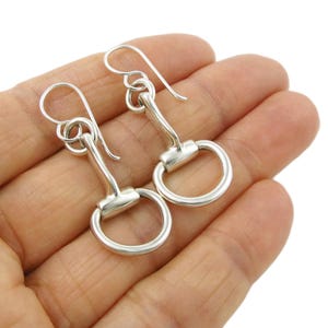 Puede incluir: Un par de pendientes plateados con forma de bocado de caballo. Cada pendiente tiene un anillo circular en la parte inferior y un gancho curvo en la parte superior para la oreja. Los pendientes se muestran sobre un fondo claro.