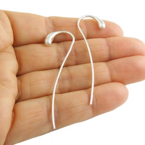 May include: A pair of silver drop earrings. Each earring has a curved, slender wire with a teardrop-shaped end. The earrings are shown against a neutral background, emphasising their minimalist design and polished finish.